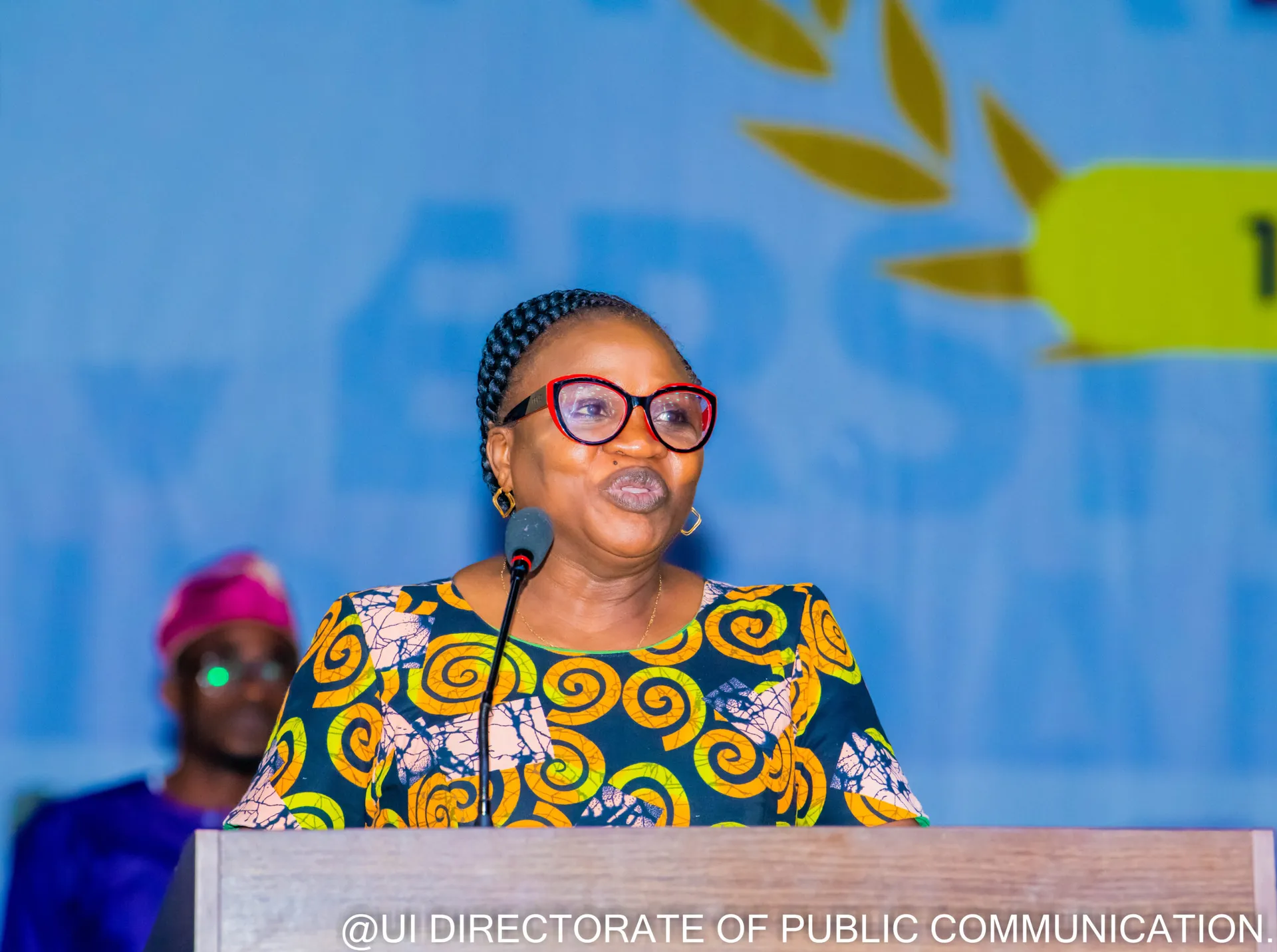 A speaker in a yellow patterned outfit addressing the forum from a lectern.