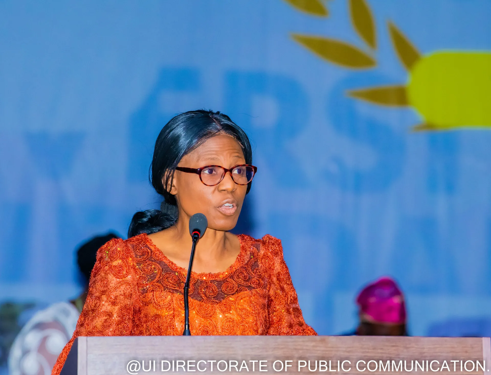 A speaker in an orange outfit addressing the forum at a lectern.